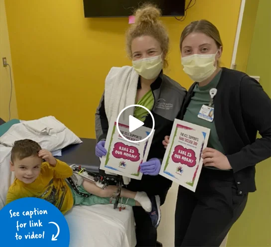 Two female physical therapists holding up Rare Disease Day signs next to a smiling young boy wearing an external fixator.