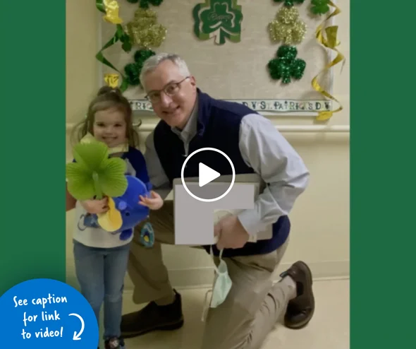 Photo of a young girl holding a big green shamrock and a toy next to Dr. Standard kneeling beside her