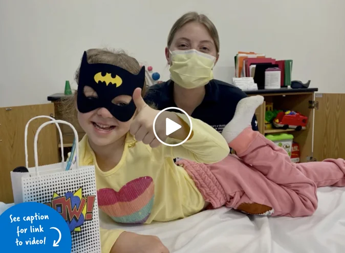 A young girl, wearing a Batman mask lying down with her thumbs up and a goody bag that says Pow! on it in front of a young woman seated behind her