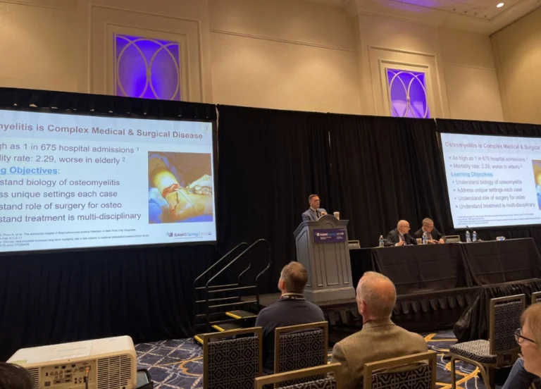 Dr. Bibbo presenting at a podium with screens showing a PowerPoint presentation in a conference room