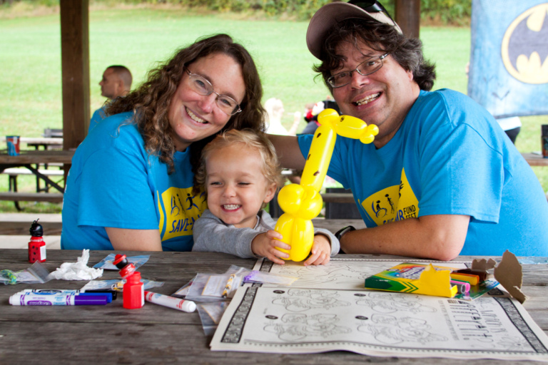 Young child holds yellow balloon animal with parents under a pavilion at Rubin Institute for Advanced Orthopedics 2017 Save-A-Limb Fund Event