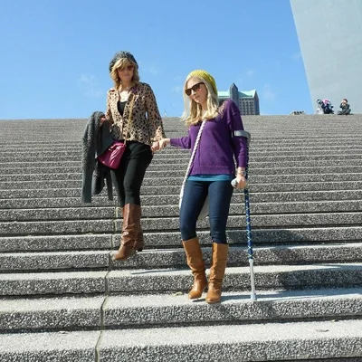 Carly as a young woman going down the stairs from the St. Louis Gateway Arch with crutches and holding a woman's hand