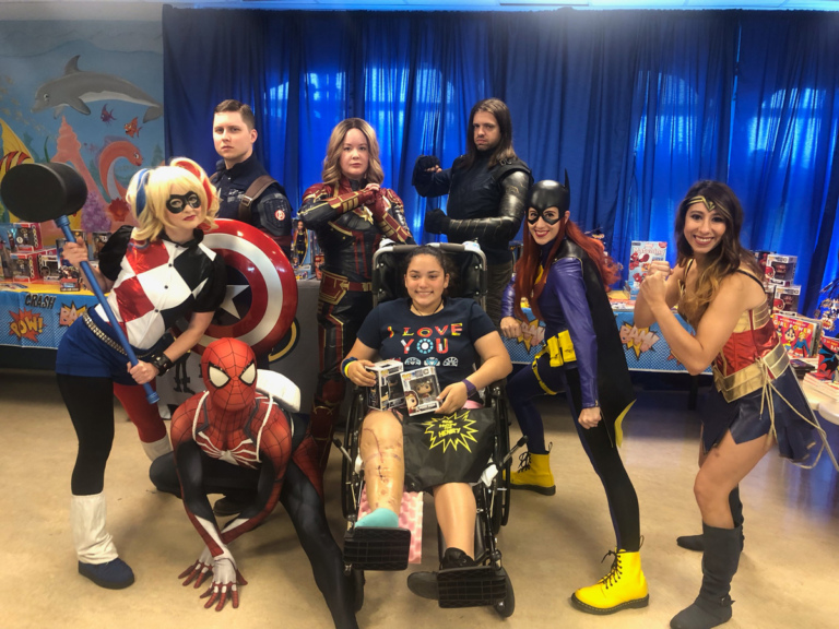 Girl patient in Wheelchair holding superhero bobbleheads posing with superheroes Wonder Woman, Bat Girl, Iron Man, Captain Marvel, Captain America and Spiderman and villain Harley Quinn