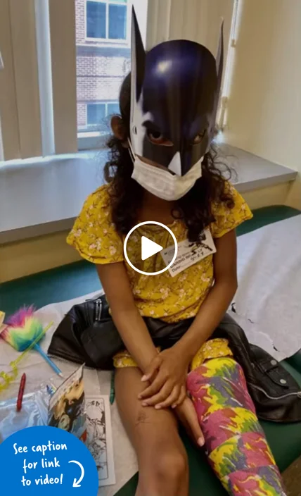 A girl sitting on an exam table with a cast on her leg and wearing a Batman mask.